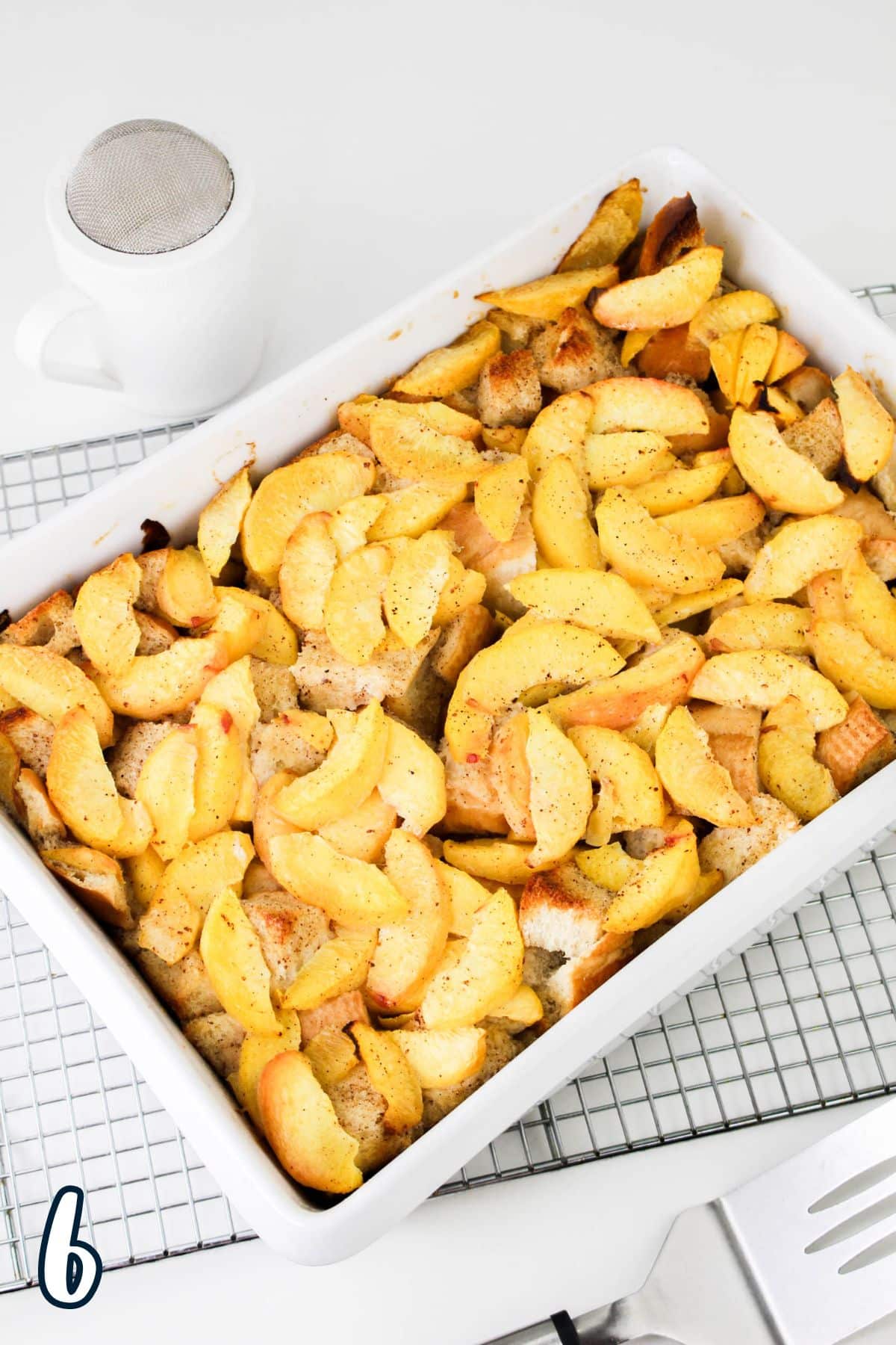 A rectangular baking dish filled with baked French toast with peaches, set on a wire rack with a white cup and a sifter.