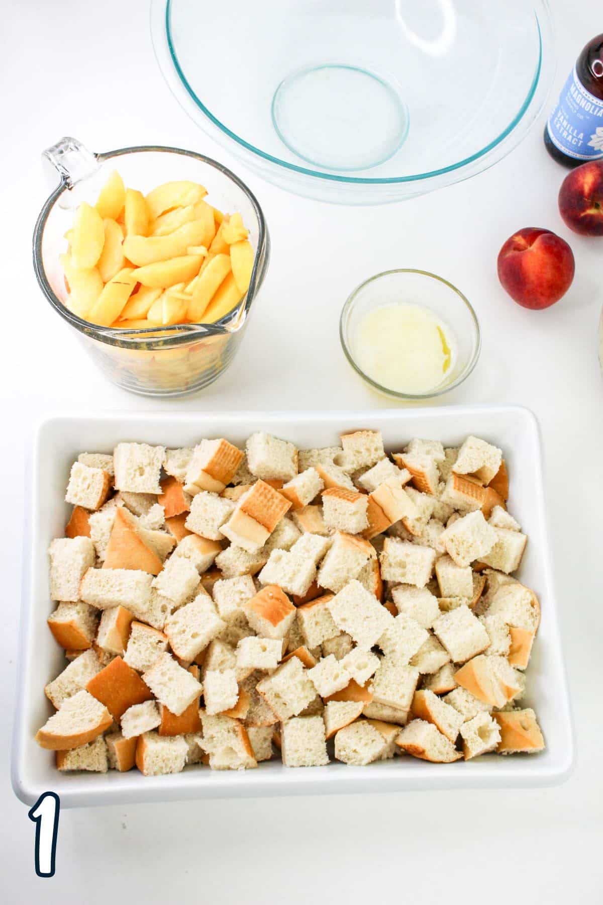 Ingredients for a dish including a white baking dish with cubed bread, a measuring cup with sliced peaches, a small bowl with melted butter, two whole peaches, and a glass mixing bowl.