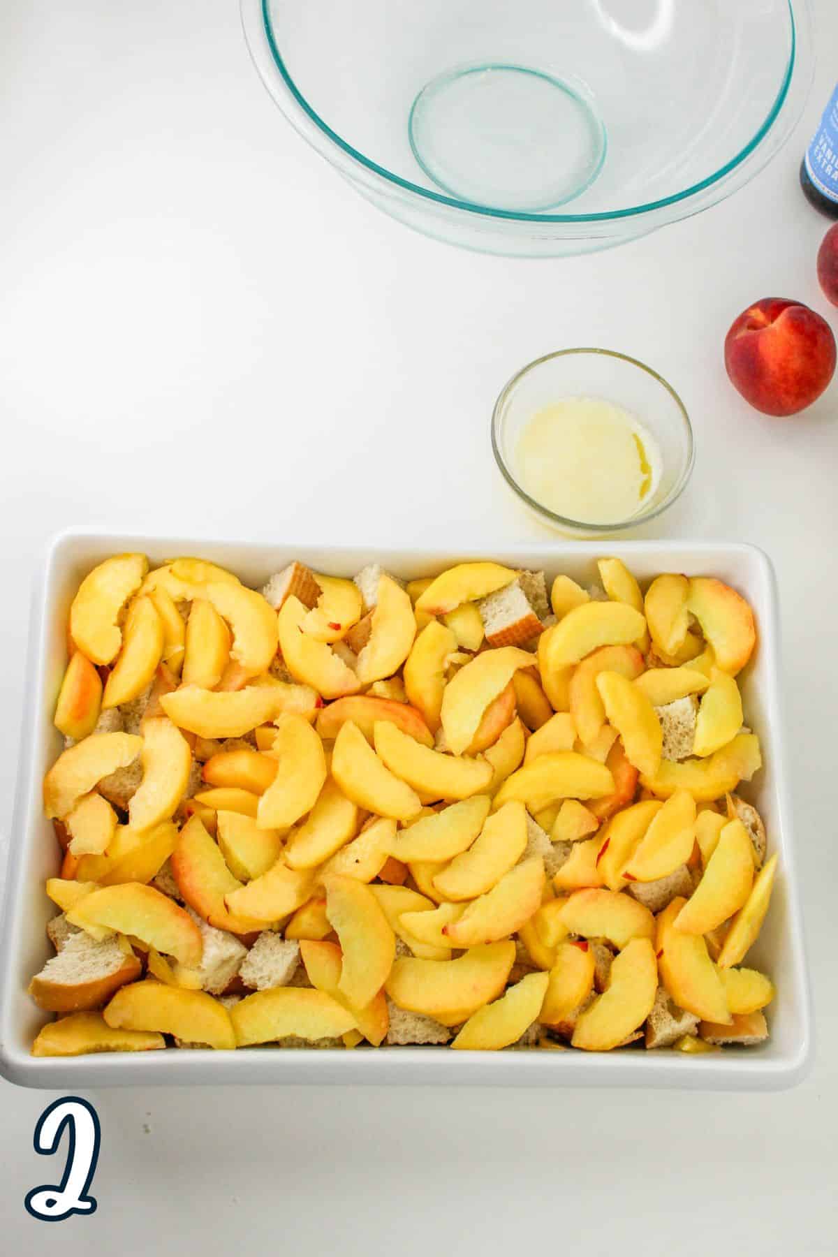 A baking dish filled with sliced peaches and pieces of bread, a small bowl of melted butter, whole peaches, and an empty glass mixing bowl are on a white surface.