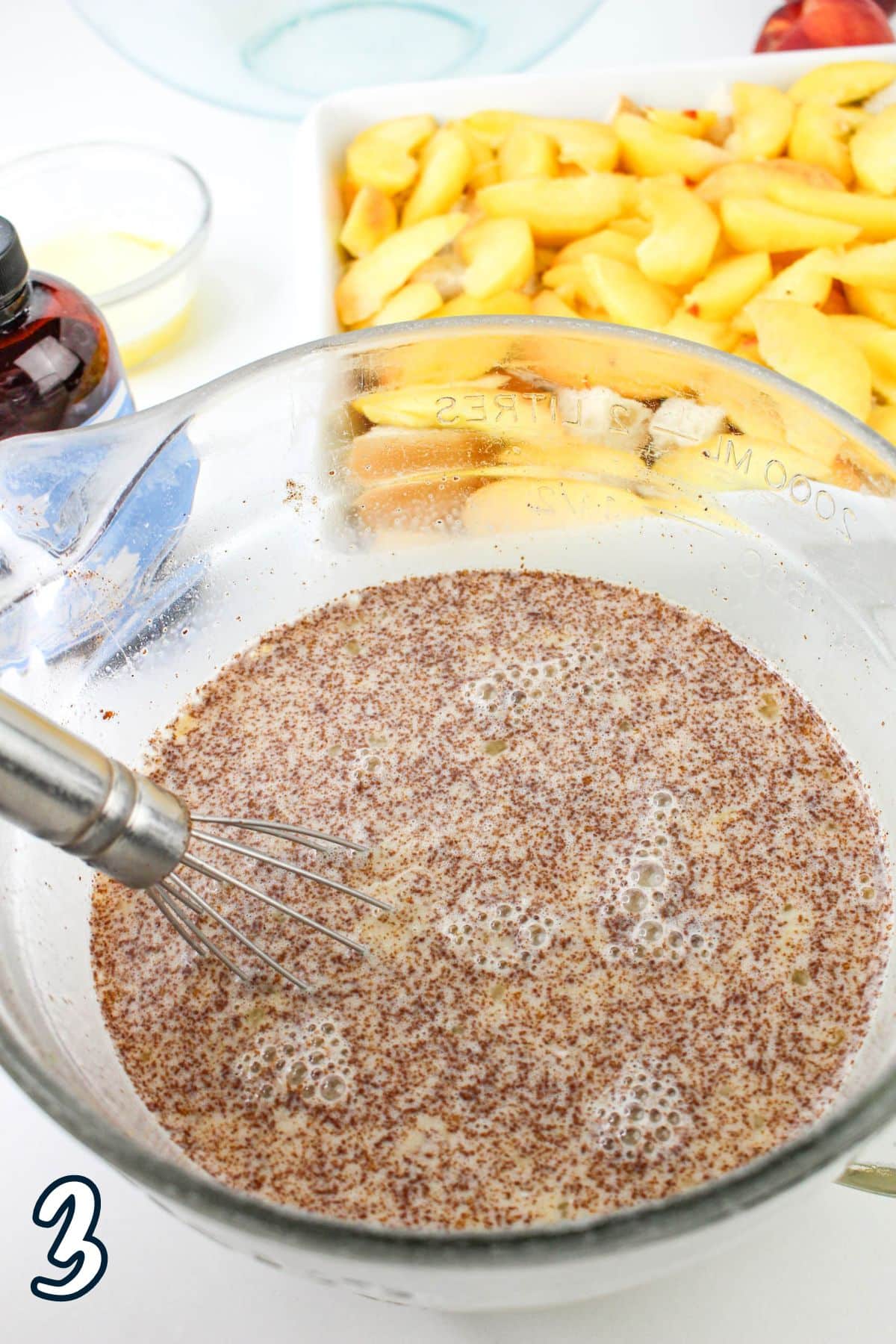 A mixing bowl containing a whisk and wet ingredients with spices. In the background, there are cut peaches and various small containers.