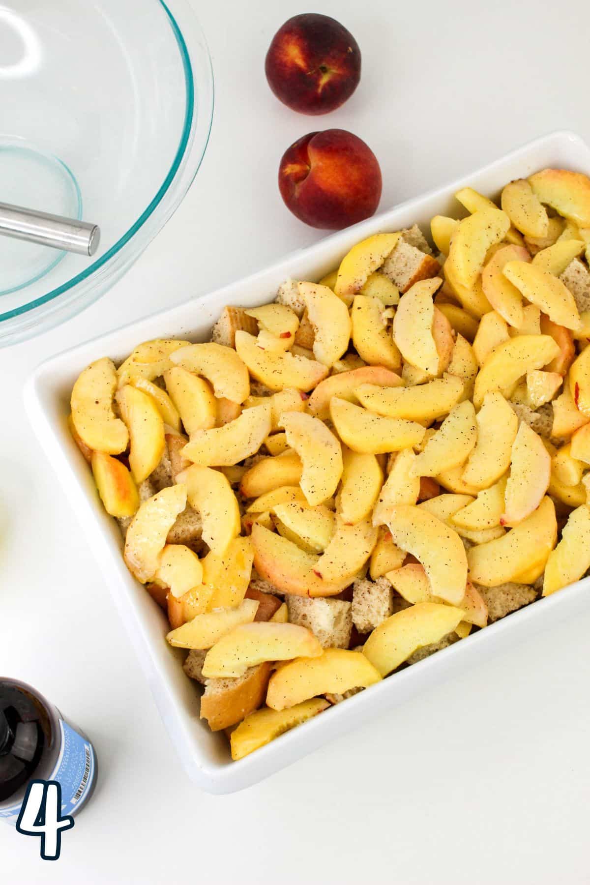 A white baking dish filled with sliced peaches on a layer of bread cubes, with whole peaches, a glass mixing bowl, a whisk, and a bottle of vanilla extract nearby. A number "4" is present in the bottom left.