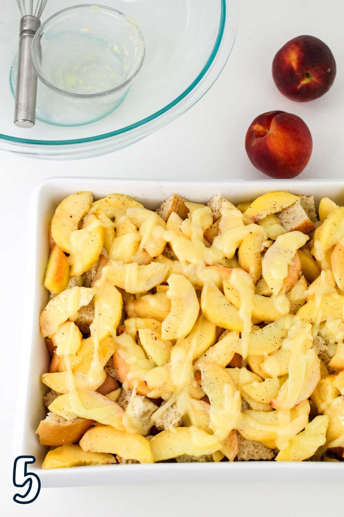 A baking dish filled with a layered mixture of sliced peaches and bread, topped with melted butter. Nearby are two whole peaches and a glass bowl with a whisk. The number 5 is in the corner.