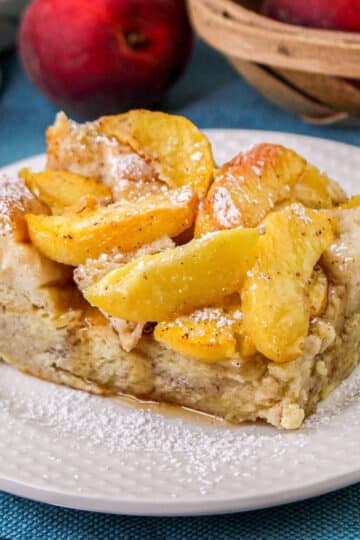 A square of peach baked French toast dusted with powdered sugar on a white plate, with fresh peaches and a basket behind it.