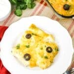 A plate with cheesy taquitos, topped with black olives and cilantro; next to the plate is a fork and rolled taquitos.
