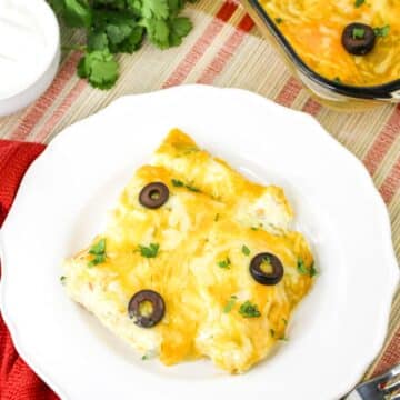 A plate with cheesy taquitos, topped with black olives and cilantro; next to the plate is a fork and rolled taquitos.