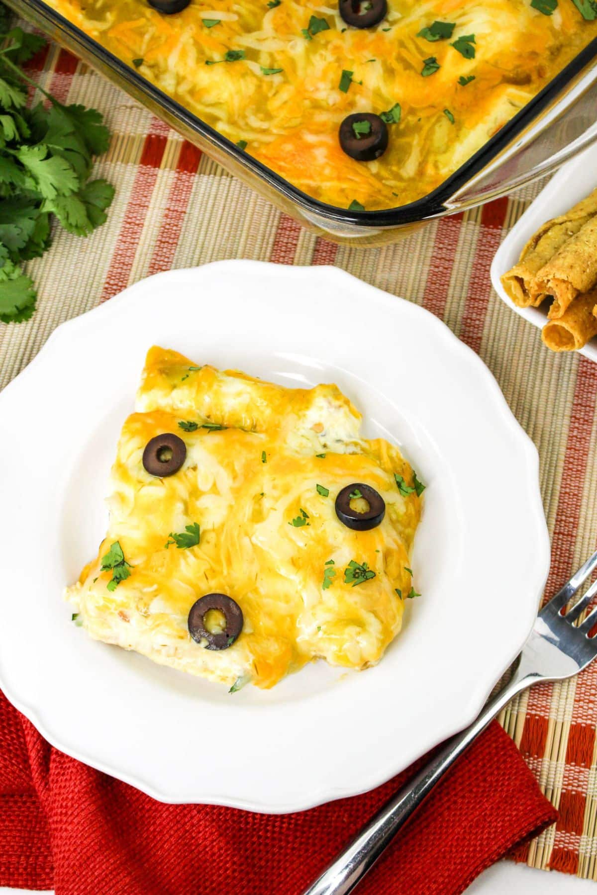 A serving of cheesy taquito enchiladas garnished with black olive slices and fresh herbs on a white plate. A casserole dish with more enchiladas and a side of fried rolls are in the background.