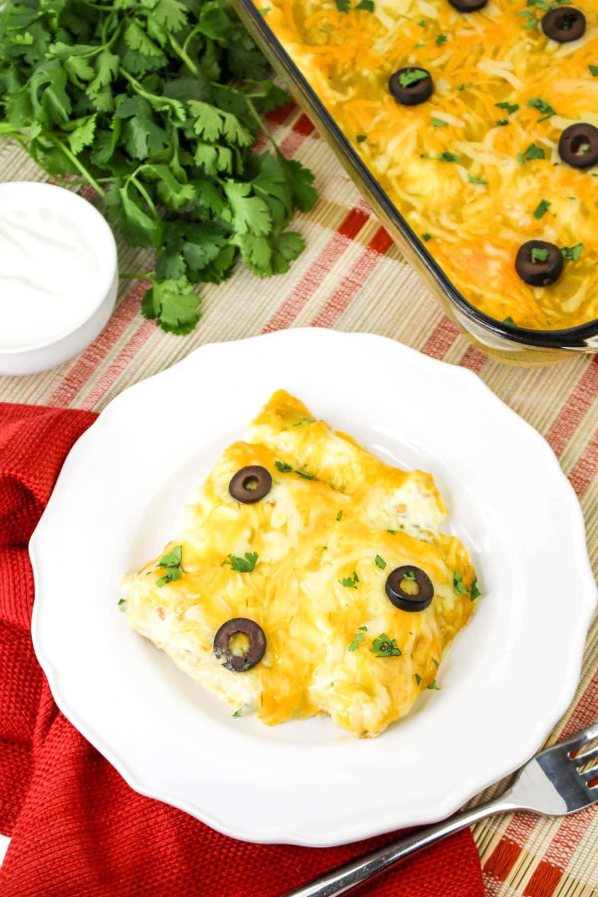 A plate of cheesy taquito casserole topped with sliced black olives and chopped herbs, set on a red and white striped tablecloth with a red napkin, and a side dish of sour cream and fresh cilantro.