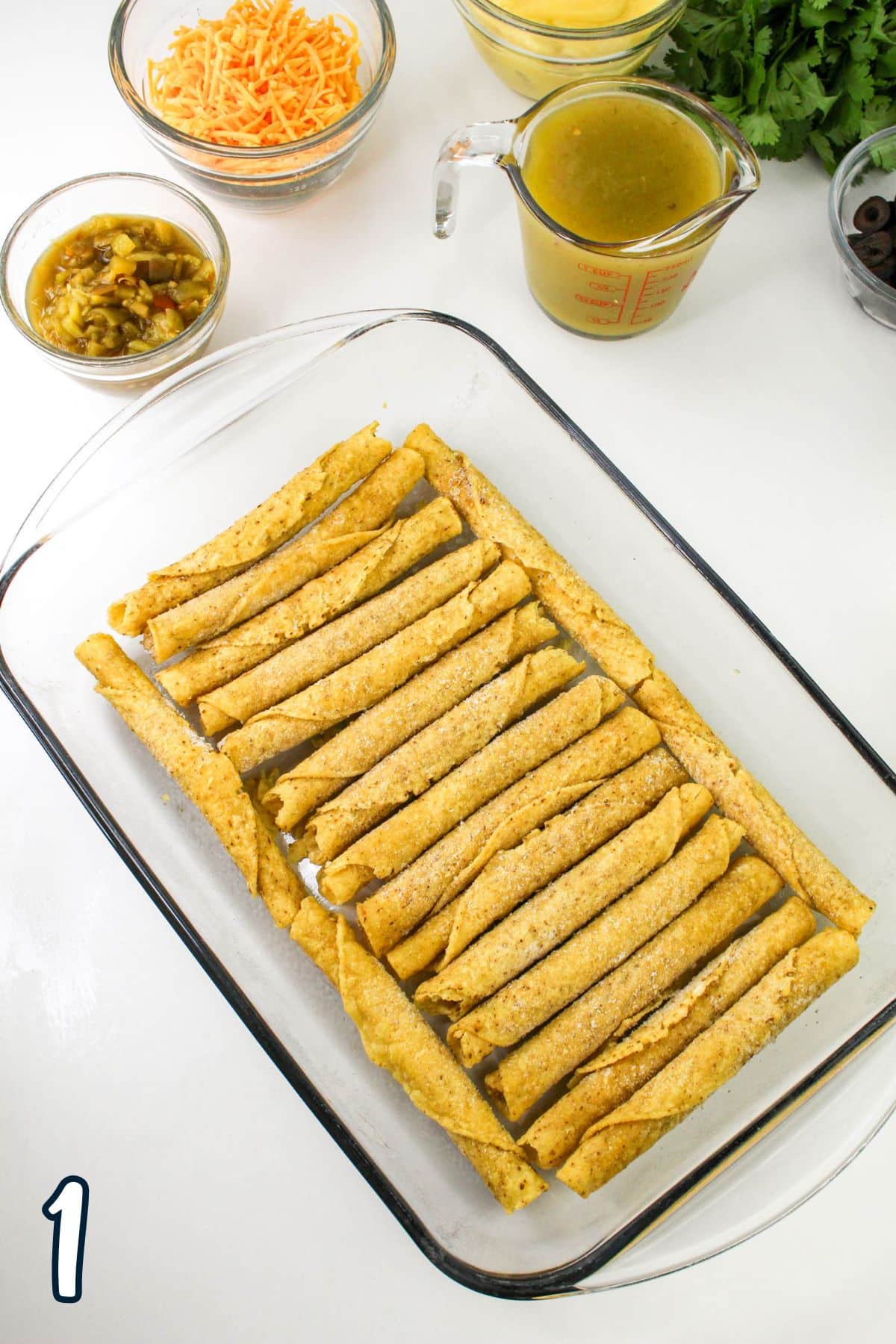 A glass baking dish filled with taquitos. Various ingredients, including cheese, sauce, olives, peppers, and cilantro, are arranged around the dish. Number "1" is visible in the corner.