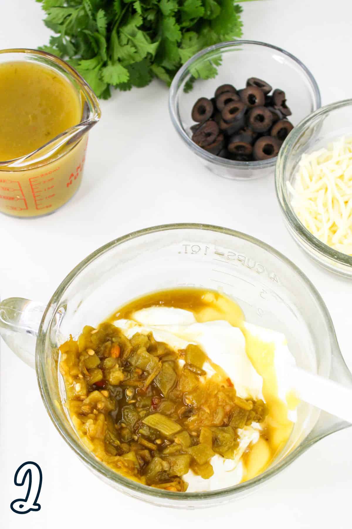 A mixing bowl containing sour cream and green chili sauce, a measuring cup with broth, a bowl of sliced black olives, and a bowl of shredded cheese, with cilantro on the side.