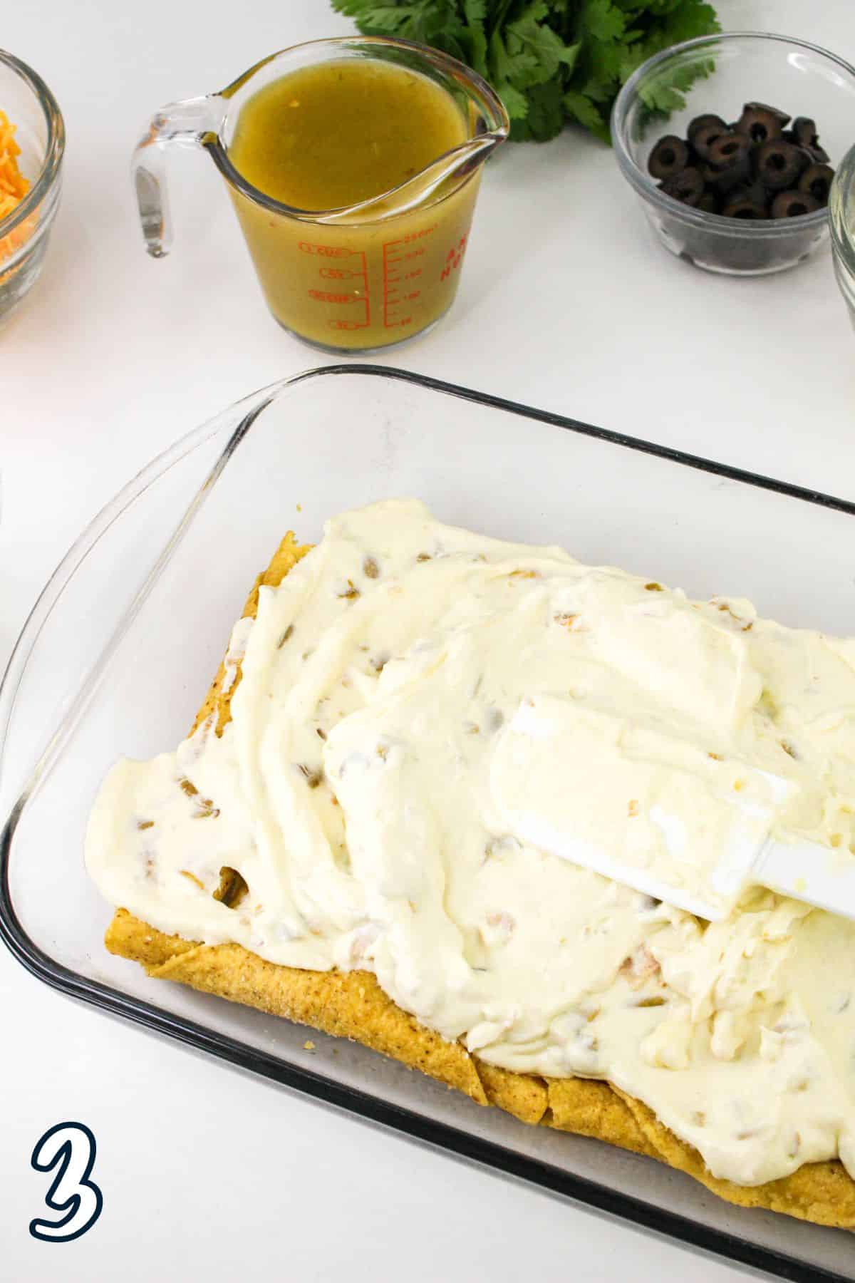 A glass baking dish with layered ingredients, including a creamy white mixture being spread with a spatula. Surrounding items include a measuring cup with sauce, a bowl of olives, shredded cheese, and greenery. The number "3" is visible in the bottom left corner.