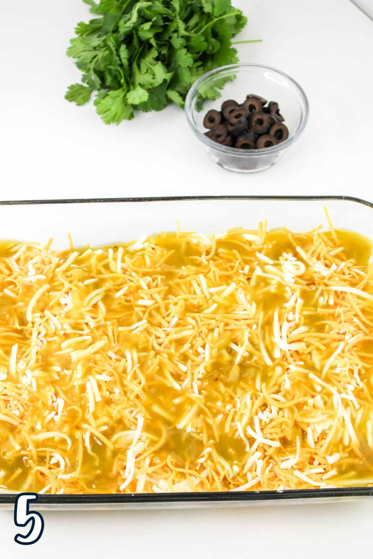 A glass baking dish filled with shredded cheese and green sauce, with a small glass bowl of sliced black olives and some fresh cilantro in the background. The number 5 is visible in the lower left corner.