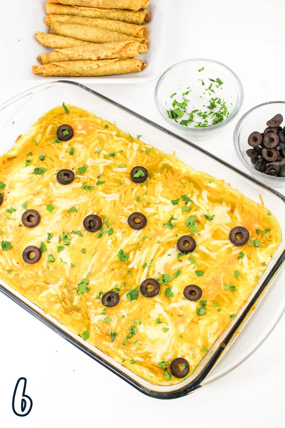 A glass baking dish with a baked casserole, topped with black olive slices and green herbs, sits on a white surface. Nearby are a bowl of chopped herbs, a bowl of black olives, and a plate of taquitos.