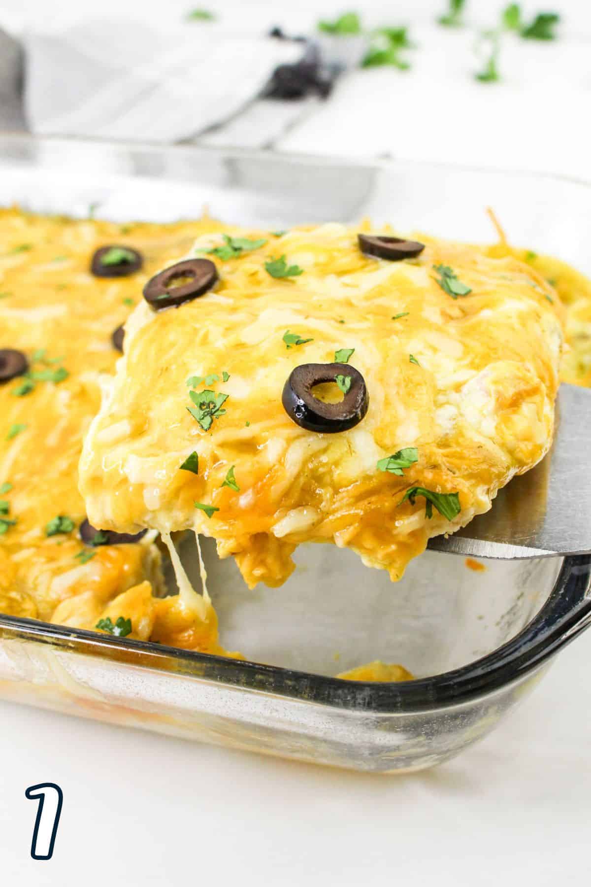 A slice of cheesy casserole with olives is being served from a glass baking dish.