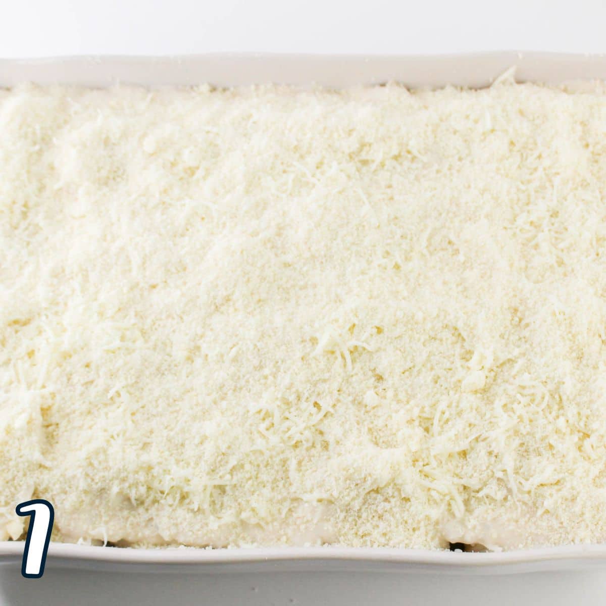 A close-up view of an uncooked, cheese-layered casserole in a white baking dish. The dish is topped with a substantial layer of grated cheese . The number "7" is seen in the bottom left corner.