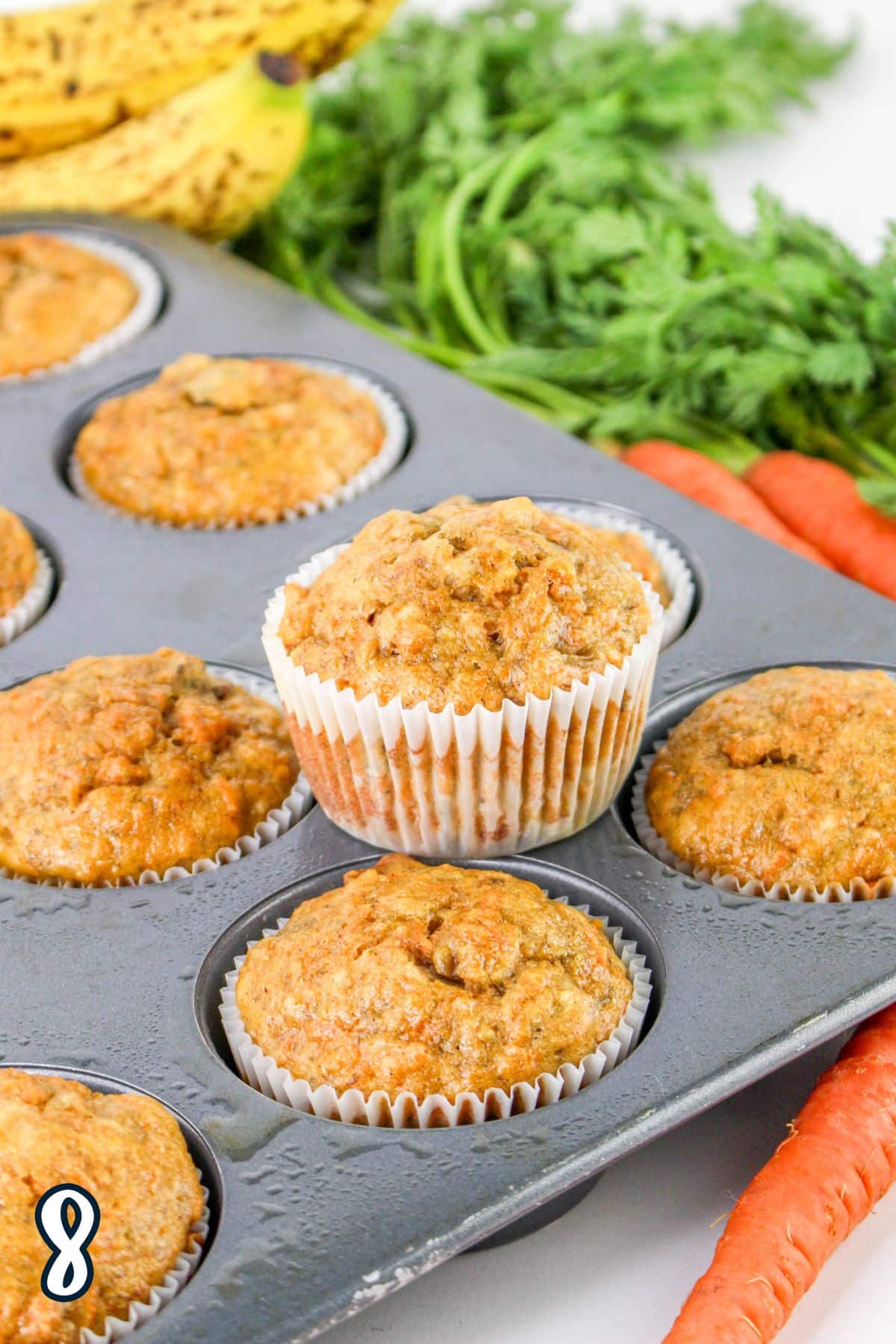 A muffin tin filled with freshly baked cupcakes, one is slightly raised, with carrots and bananas in the background.