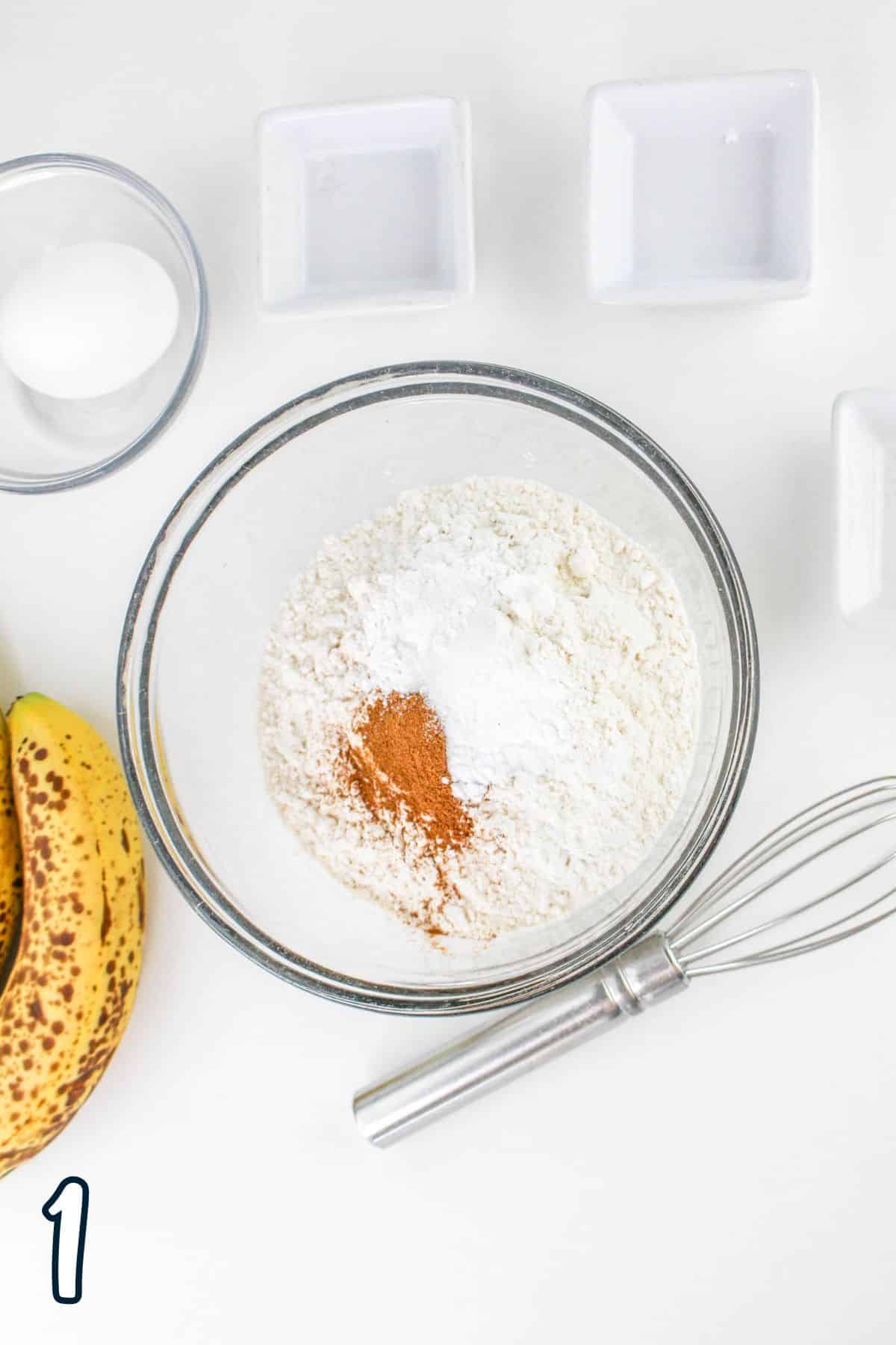 Bowl with flour and spices, whisk, banana, and an egg on a white surface, with number "1" at the bottom left corner.