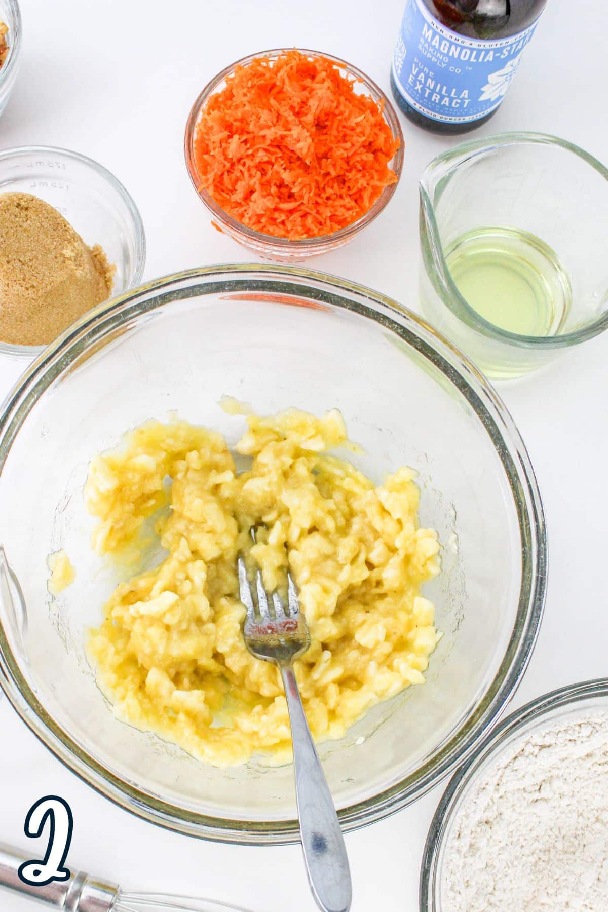 Mashed bananas in a bowl with surrounding ingredients including grated carrots, vanilla extract, oil, brown sugar, and flour.