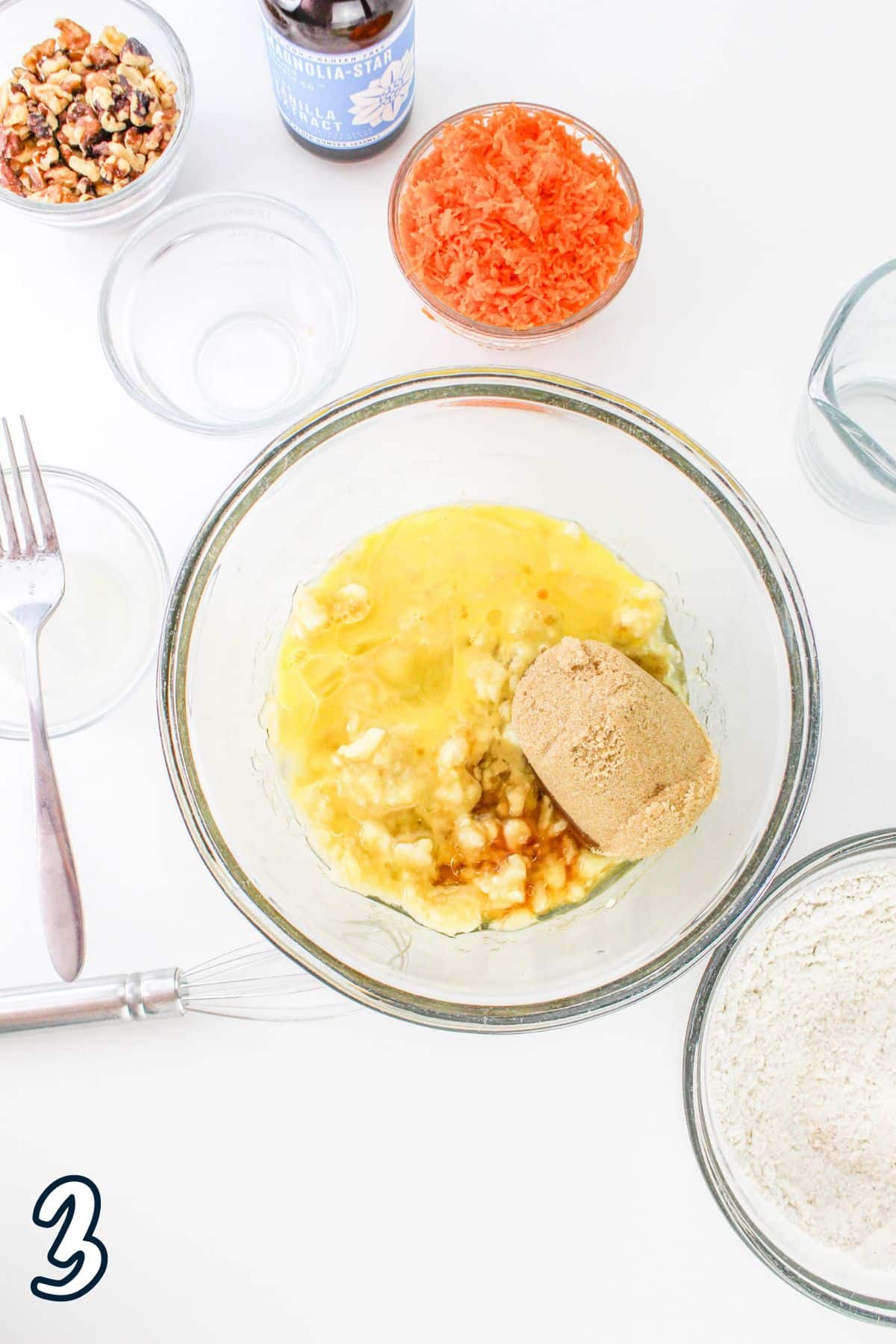 Ingredients, including eggs, brown sugar, and vanilla extract, in a glass bowl. Grated carrots and nuts are also shown.