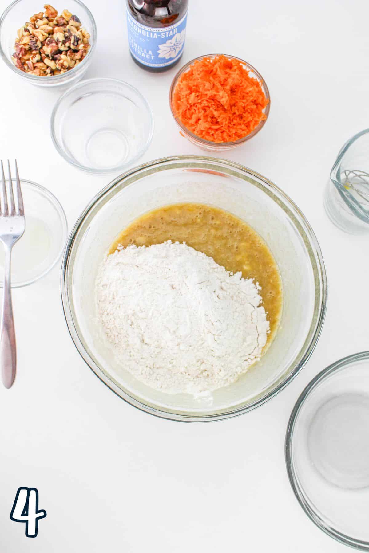 Bowl with flour and wet mixture surrounded by grated carrots, chopped nuts, vanilla extract, and empty bowls. Fork on the left.