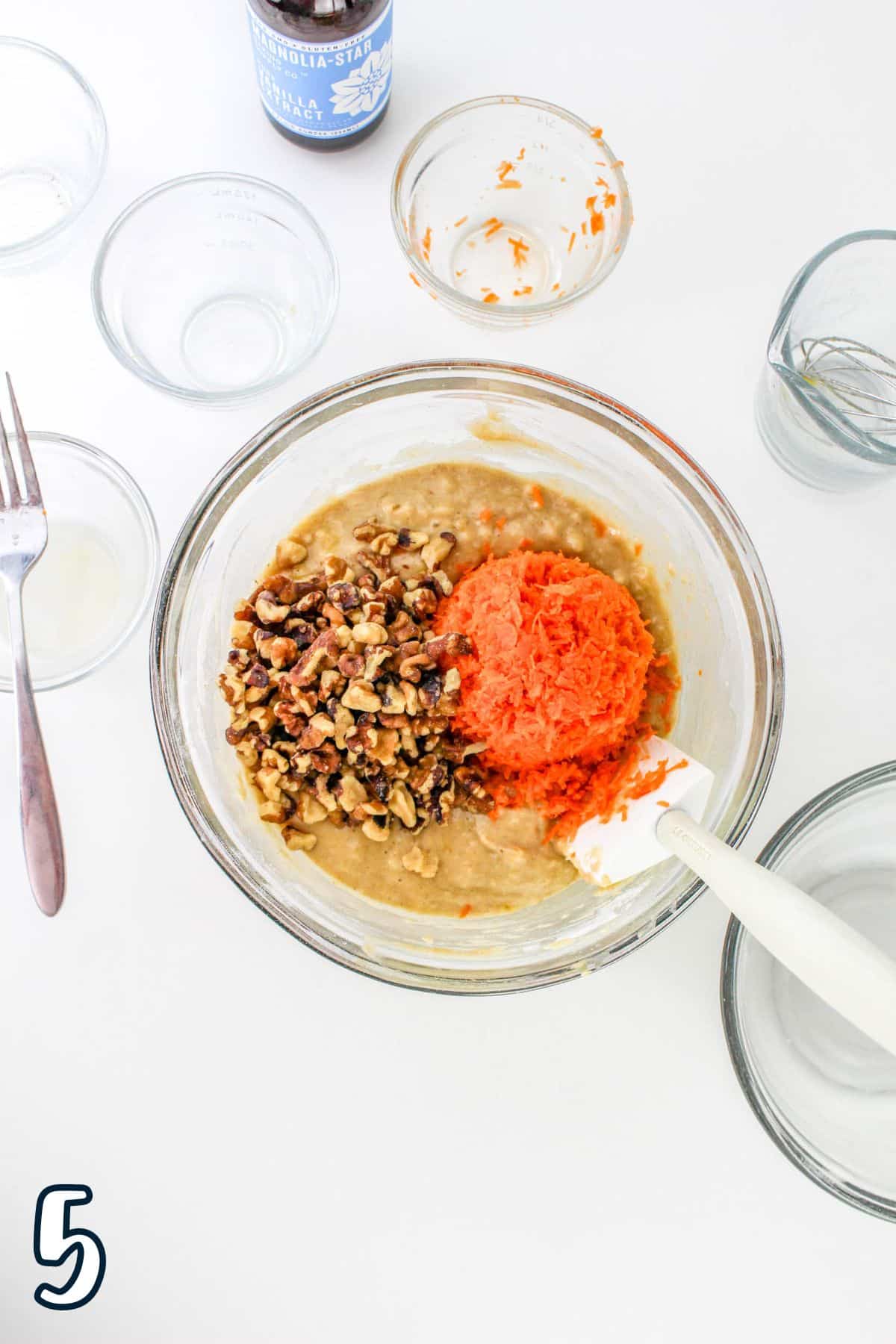 Mixing bowl containing a batter with shredded carrots and chopped nuts, surrounded by empty bowls and utensils. Number 5 in corner.