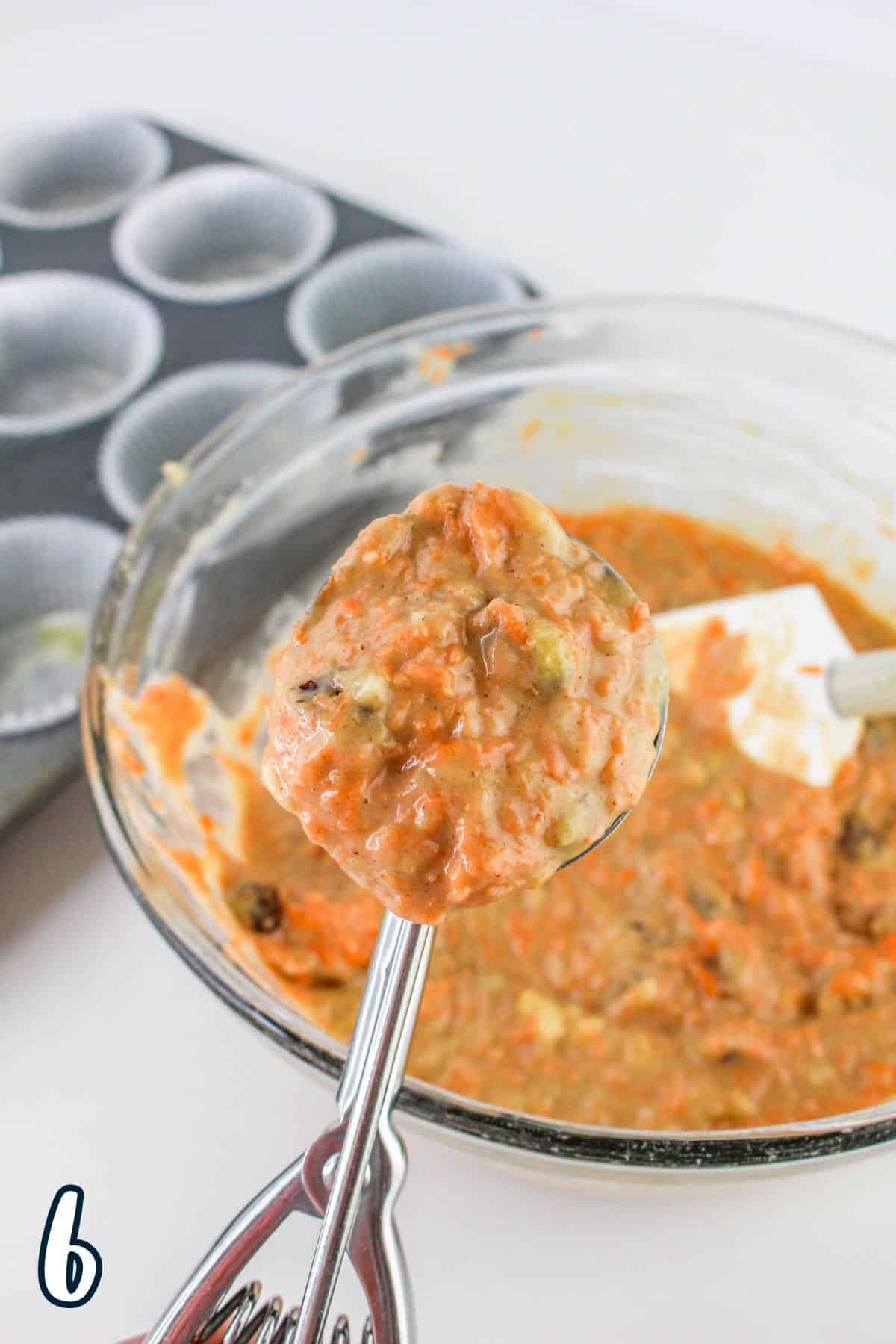 A scoop of muffin batter held above a bowl, with an empty muffin tin and liners in the background.