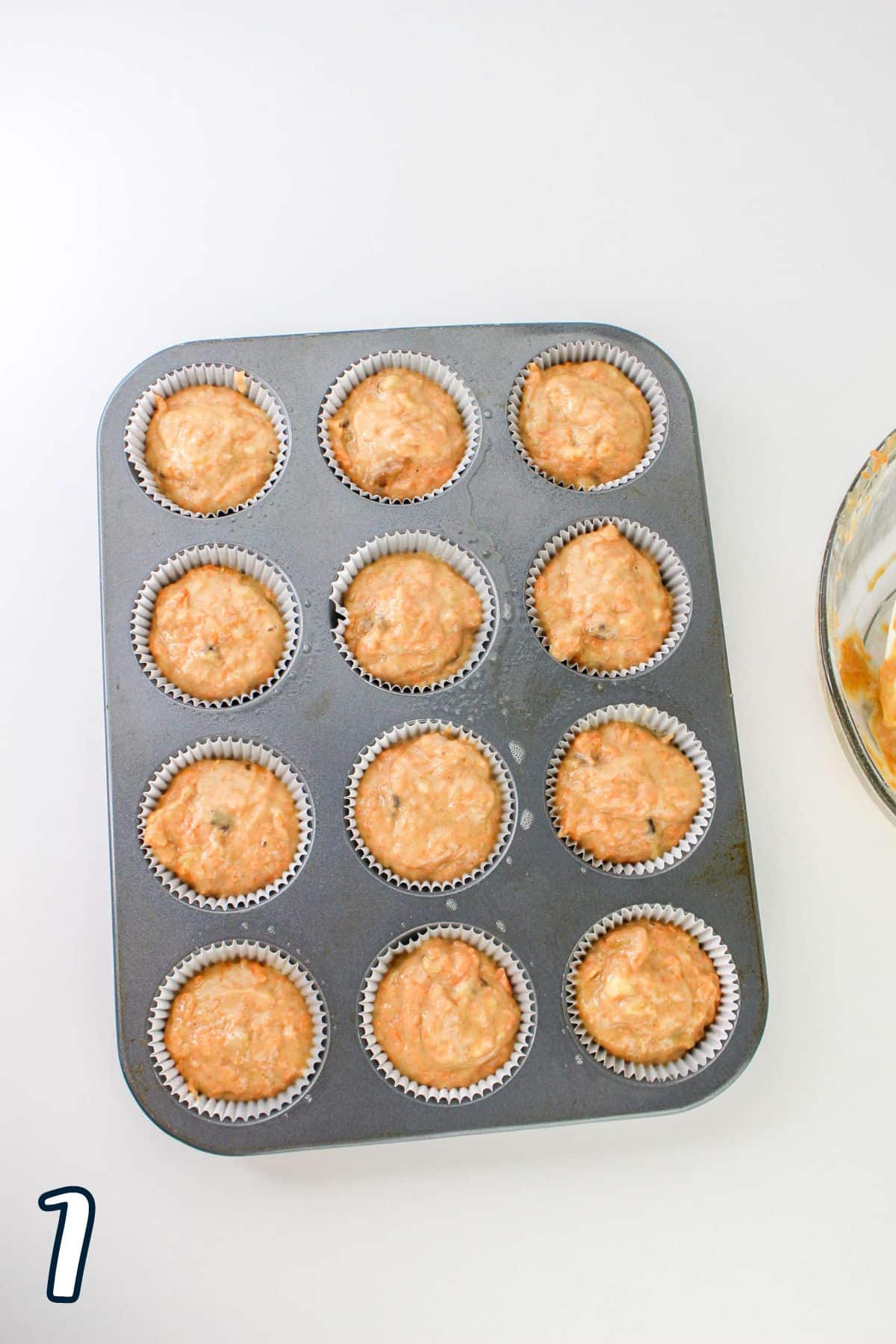 A muffin tin with 12 unbaked muffin batter portions, on a white surface. The number 1 is in the bottom left corner.