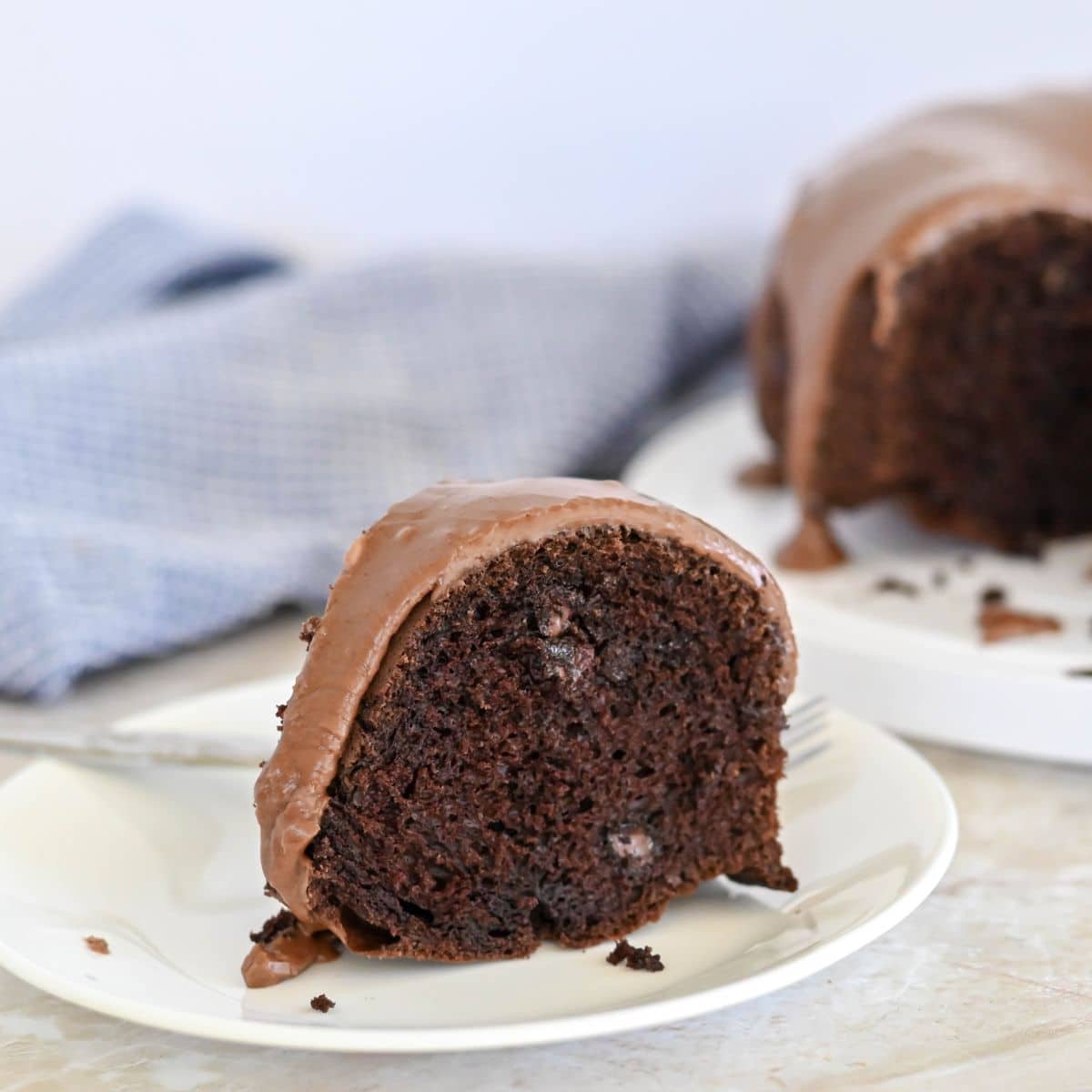 A slice of chocolate Bundt cake with chocolate icing on a white plate, with a blue cloth in the background.