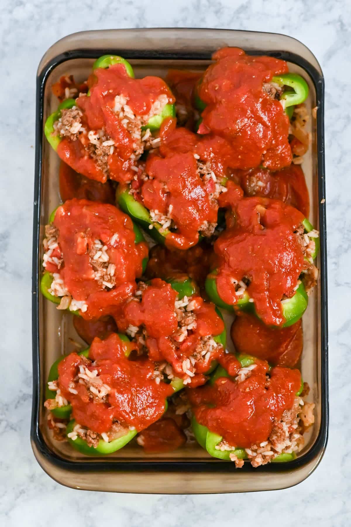 A tray of baked green bell peppers stuffed with rice and ground meat, topped with red sauce.