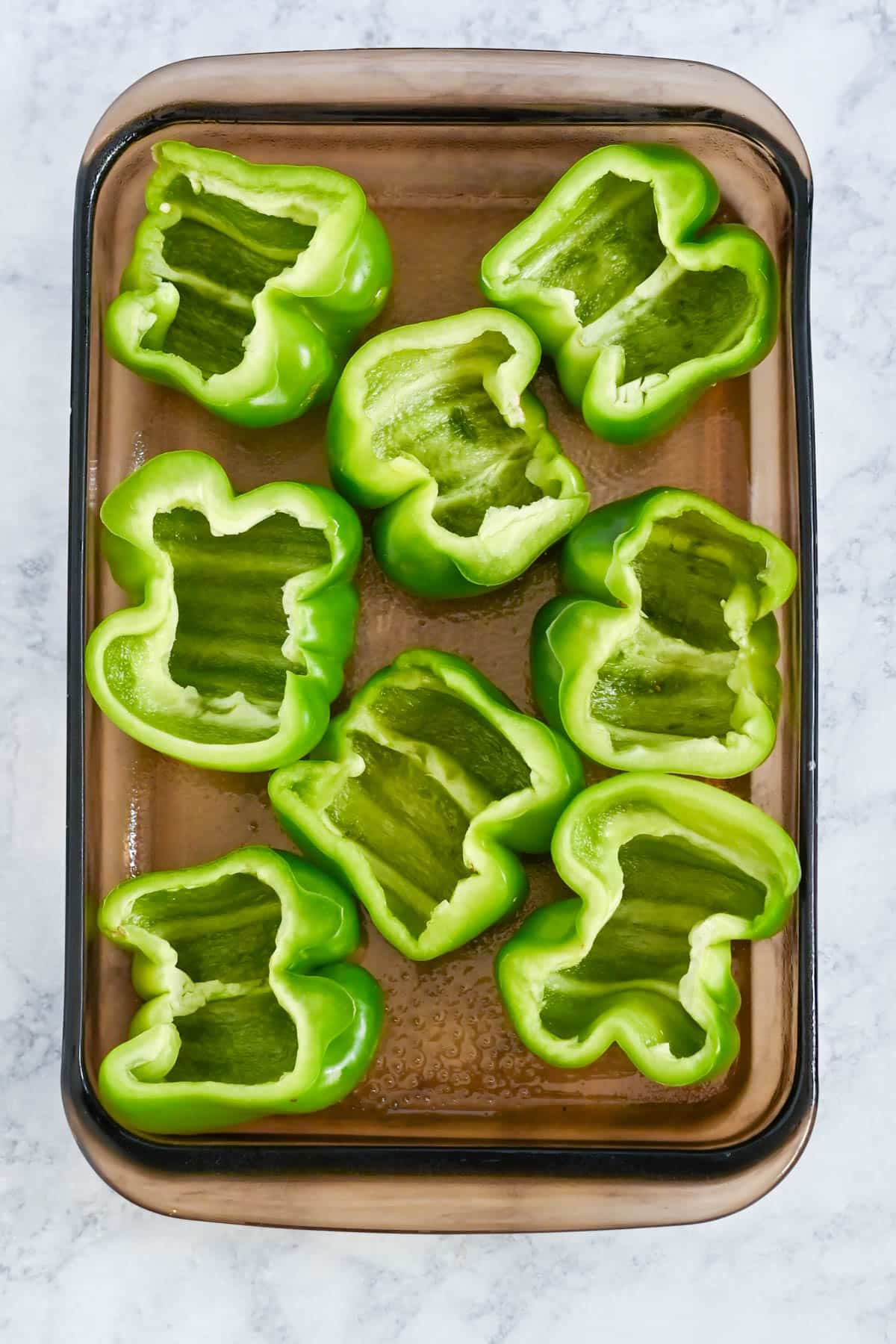 Casserole dish with eight green bell peppers cut in half, placed cut-side up on a marble counter.