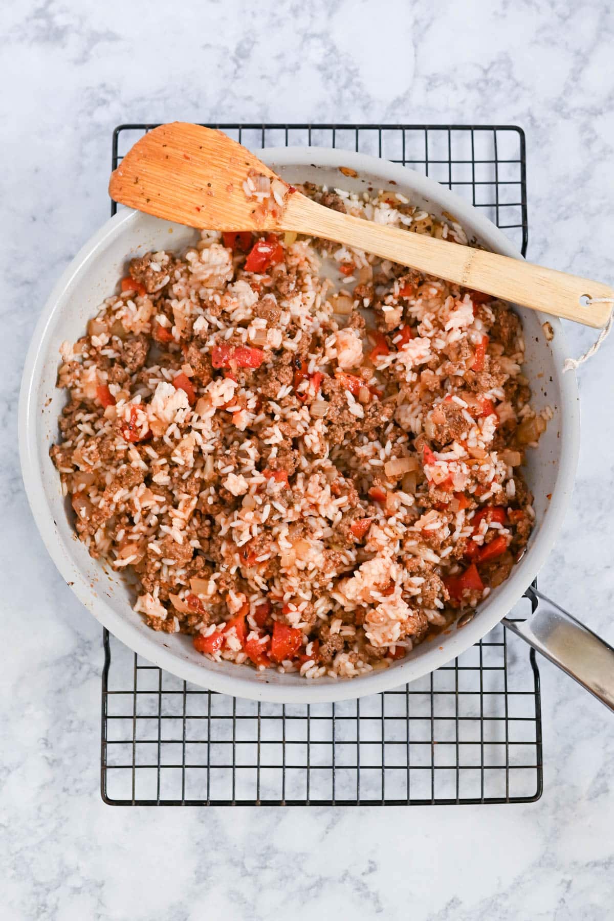 A skillet filled with rice, ground meat, tomatoes, and spices, placed on a cooling rack with a wooden spatula.