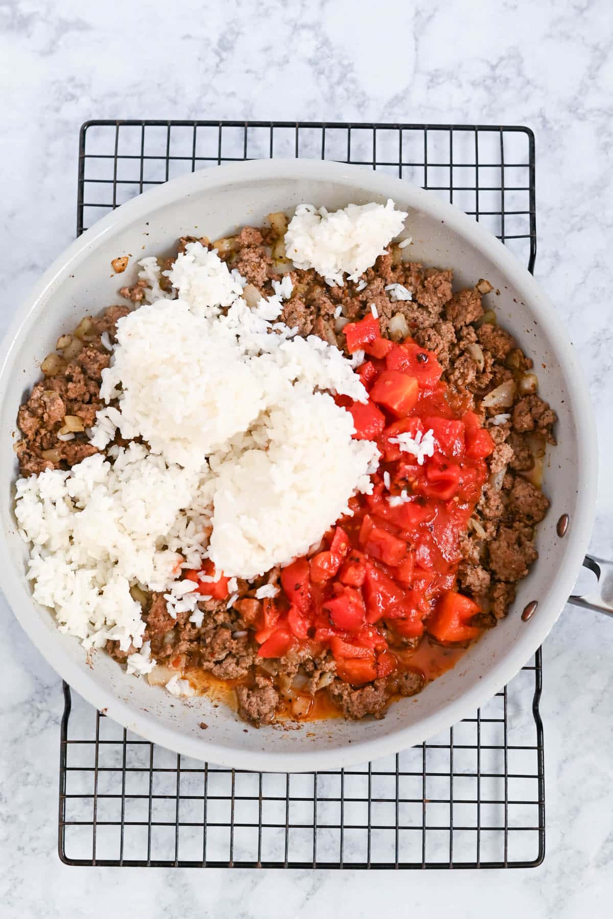 A skillet on a cooling rack contains ground meat, diced tomatoes, and a mound of rice.