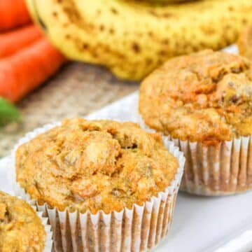 Close-up of three carrot muffins in white paper liners, with blurred bananas and carrots in the background.