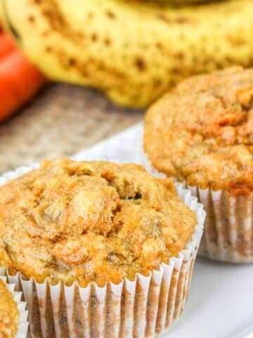 Close-up of three carrot muffins in white paper liners, with blurred bananas and carrots in the background.