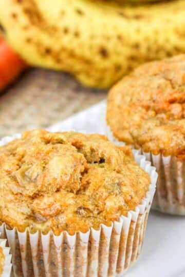 Close-up of three carrot muffins in white paper liners, with blurred bananas and carrots in the background.