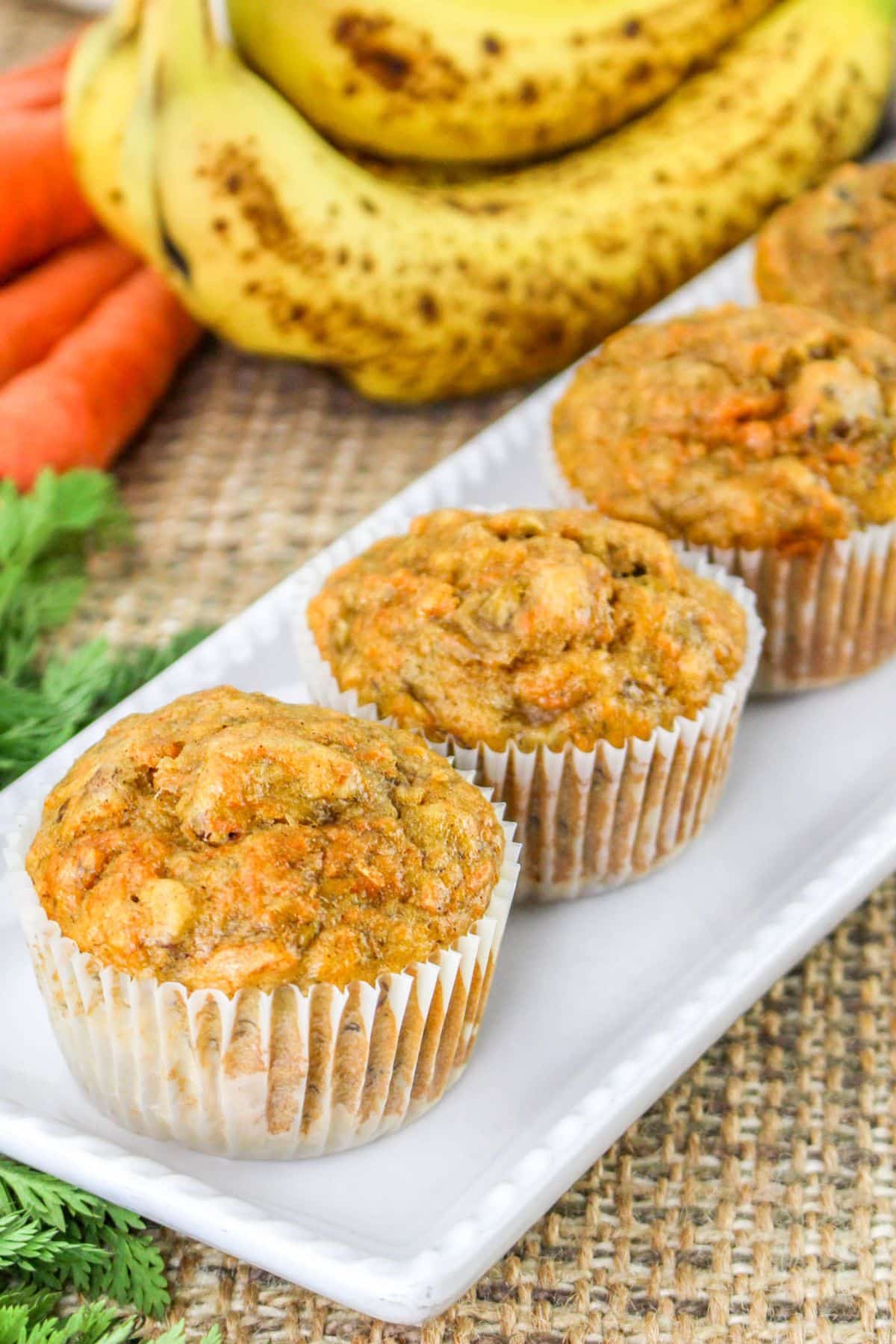 Three carrot muffins on a white plate with ripe bananas and carrots in the background.