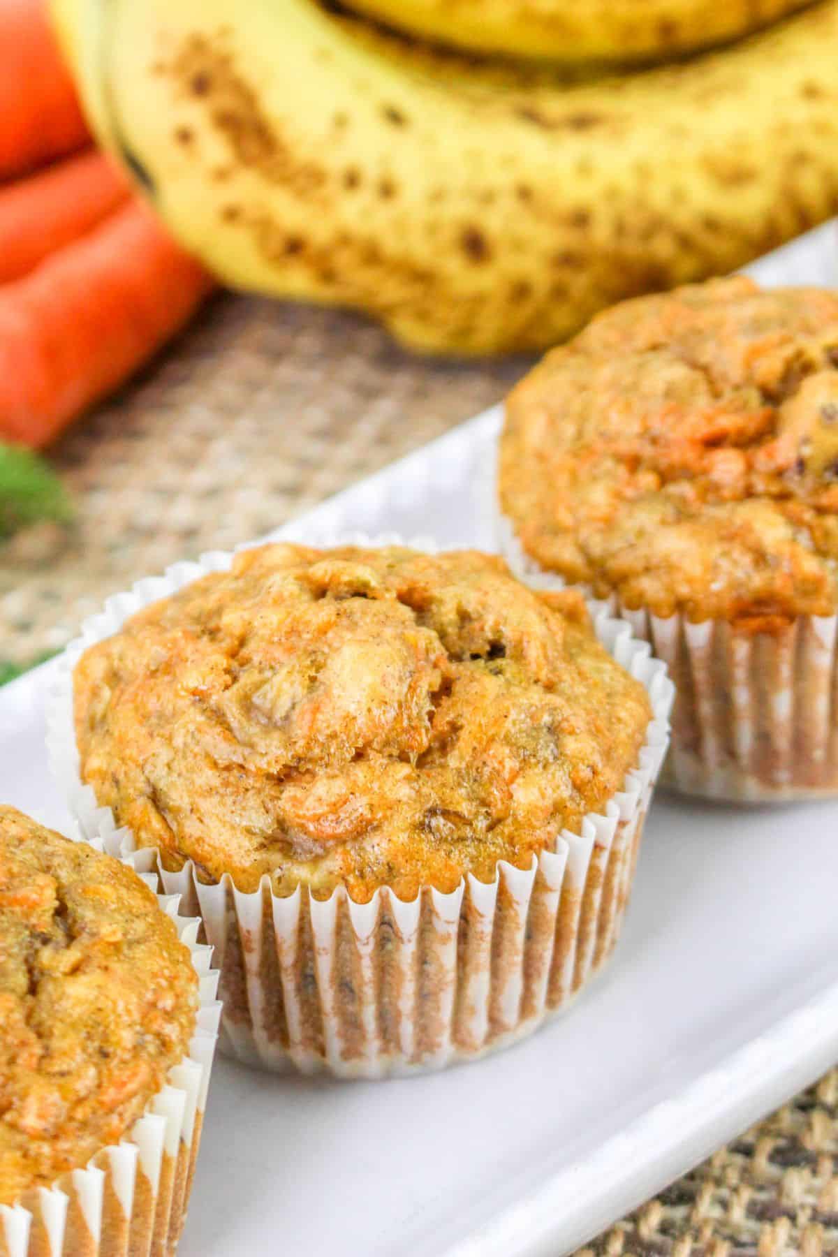 Close-up of three banana carrot muffins on a white plate, with ripe bananas and carrots in the background.