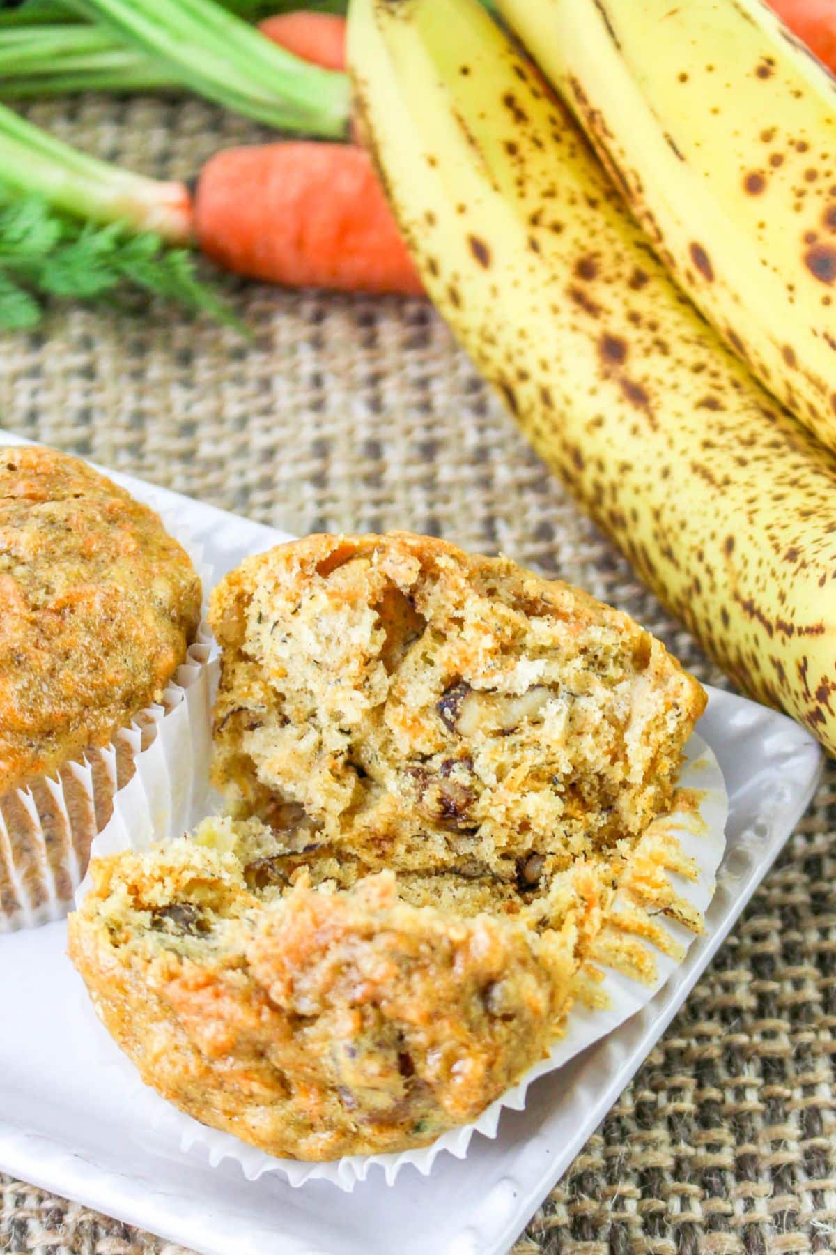 Carrot banana muffin broken in half, with bananas and carrots in the background on a burlap surface.