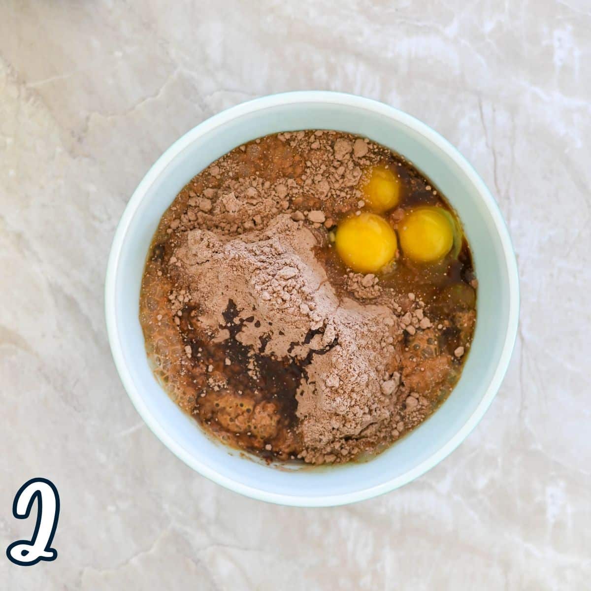 Bowl with cake mix, eggs, and cocoa powder on a marble surface.