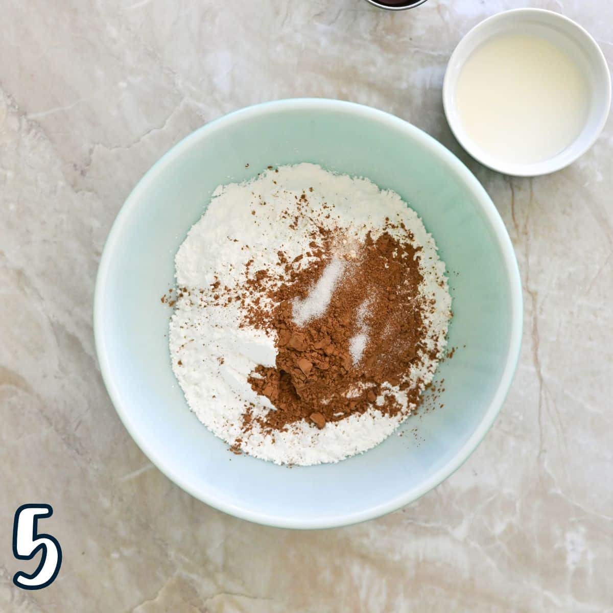 Bowl with powdered sugar, cocoa powder, and sugar on a marble surface; a small cup of milk beside it.