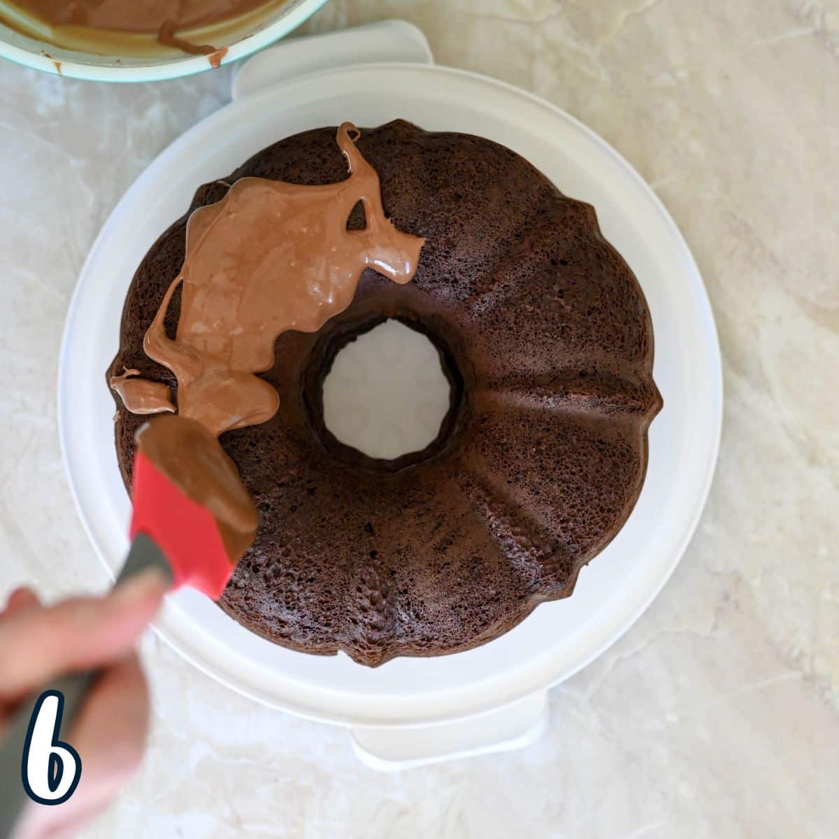 Person spreading chocolate glaze on a bundt cake with a red spatula.