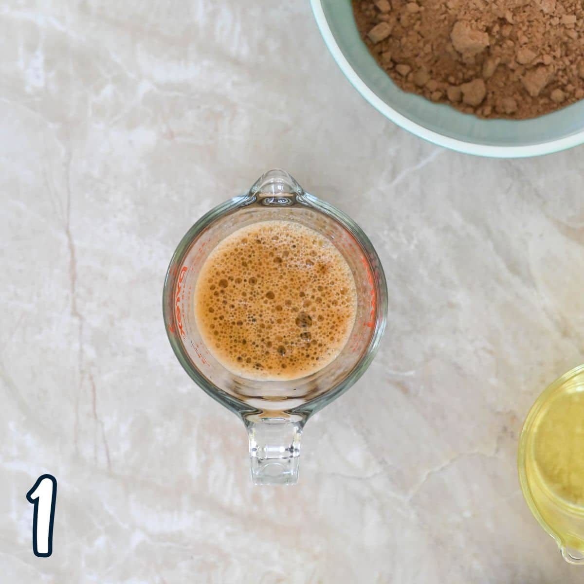 A measuring cup with a brown frothy liquid next to a bowl of cocoa powder and a bowl of oil on a marble surface.