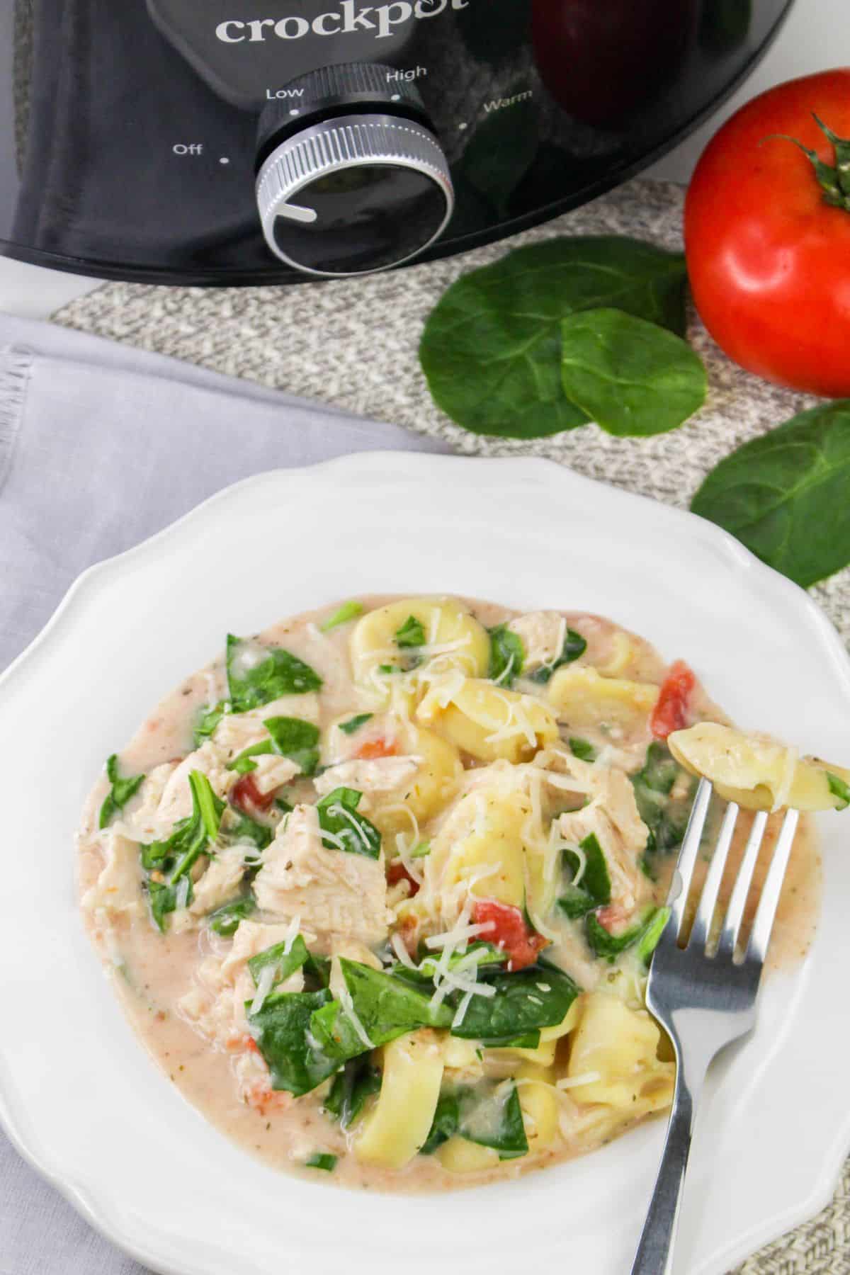 Plate of creamy chicken tortellini with spinach and tomatoes, served in front of a slow cooker. A fork rests on the plate.