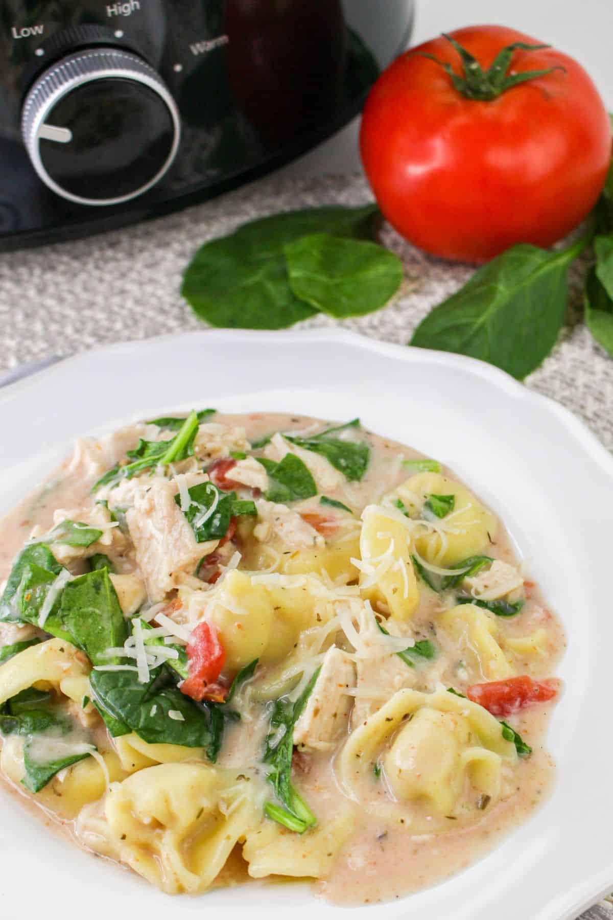 A plate of creamy tortellini with spinach and diced tomatoes, with fresh spinach leaves and a tomato beside a slow cooker.