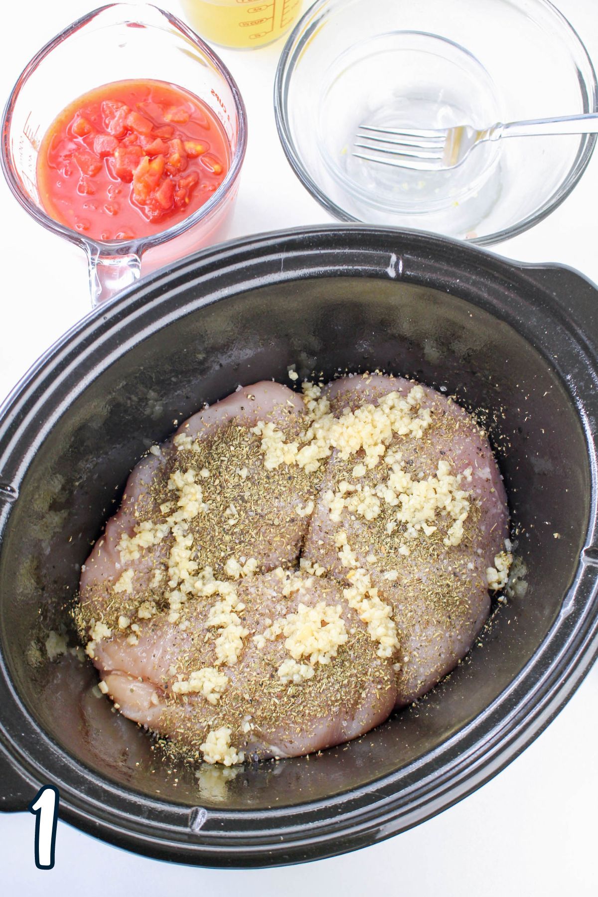 Raw chicken breasts with seasoning and minced garlic in a slow cooker pot, next to a bowl of diced tomatoes and a glass bowl.