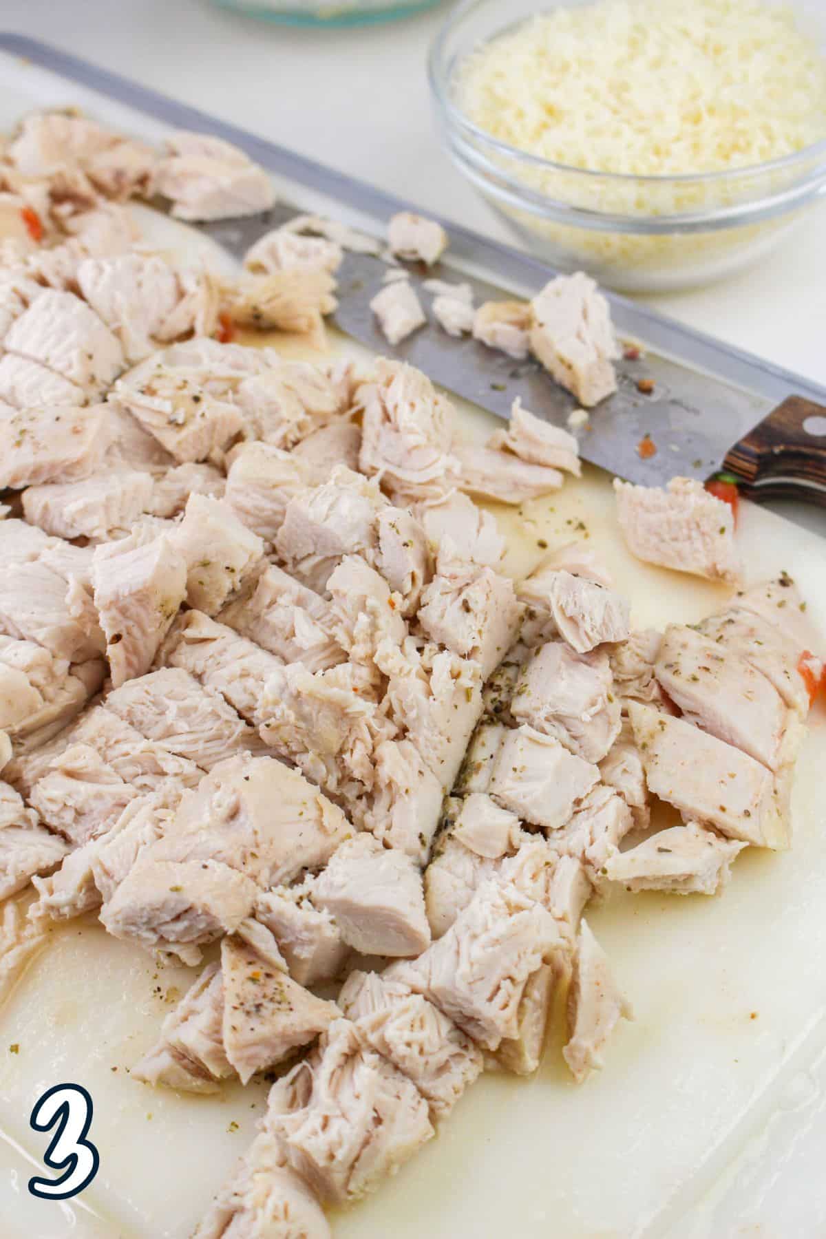 Chopped cooked chicken on a cutting board with a knife, a bowl of shredded cheese in the background, and the number '3' in the corner.