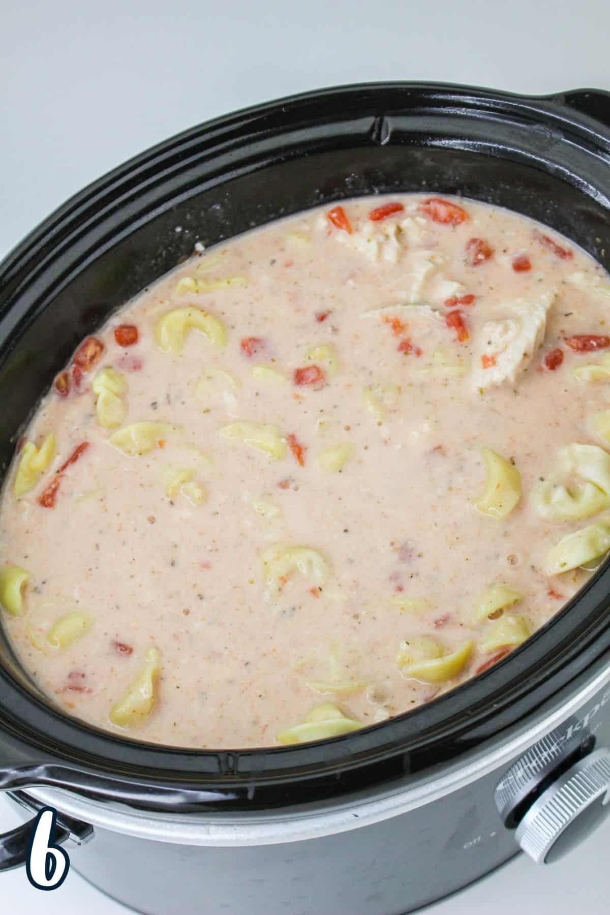 A slow cooker filled with a creamy soup containing tortellini, diced tomatoes, and chunks of chicken.