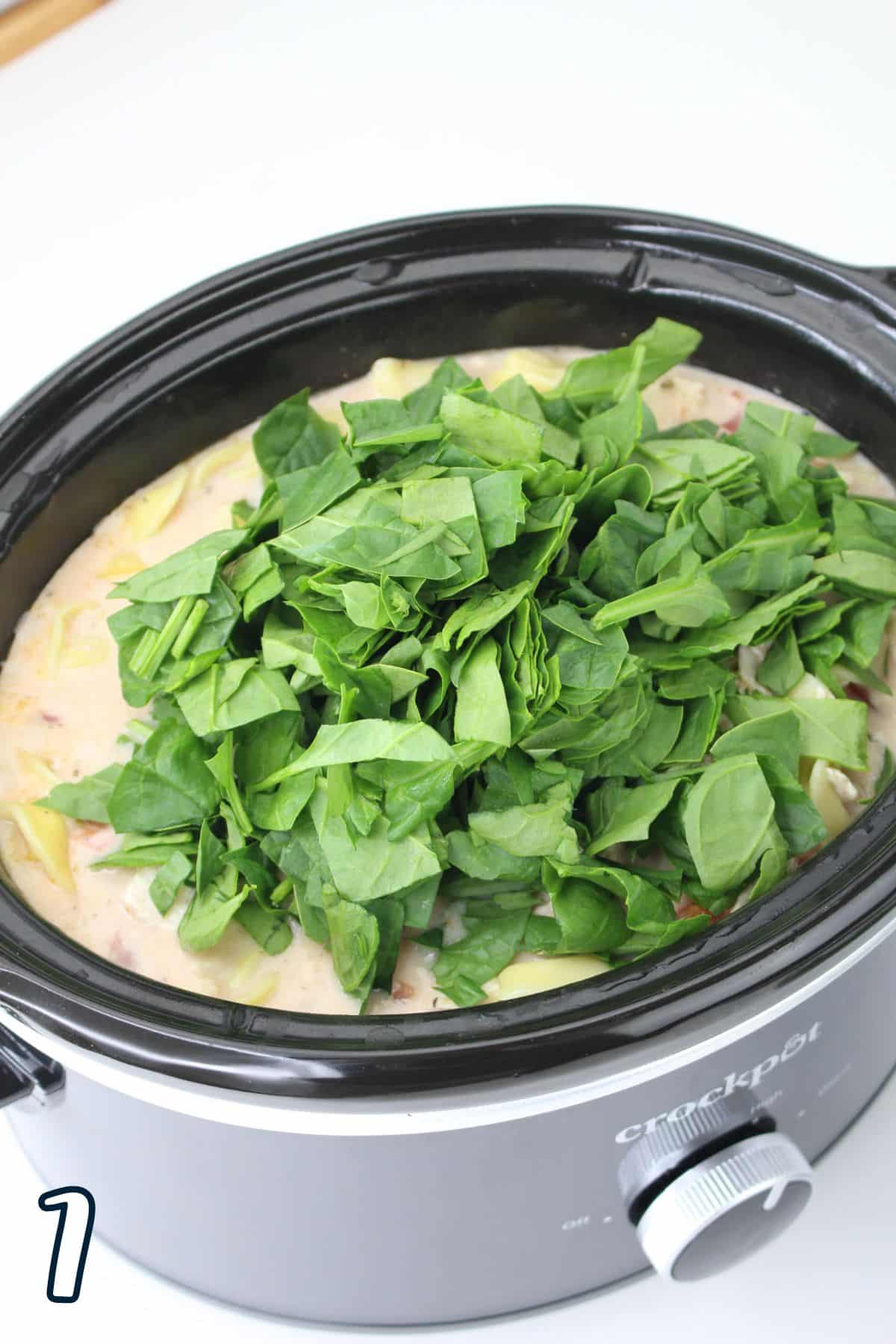 A slow cooker filled with a creamy dish topped with fresh, chopped spinach. A dial and a number "1" are visible.