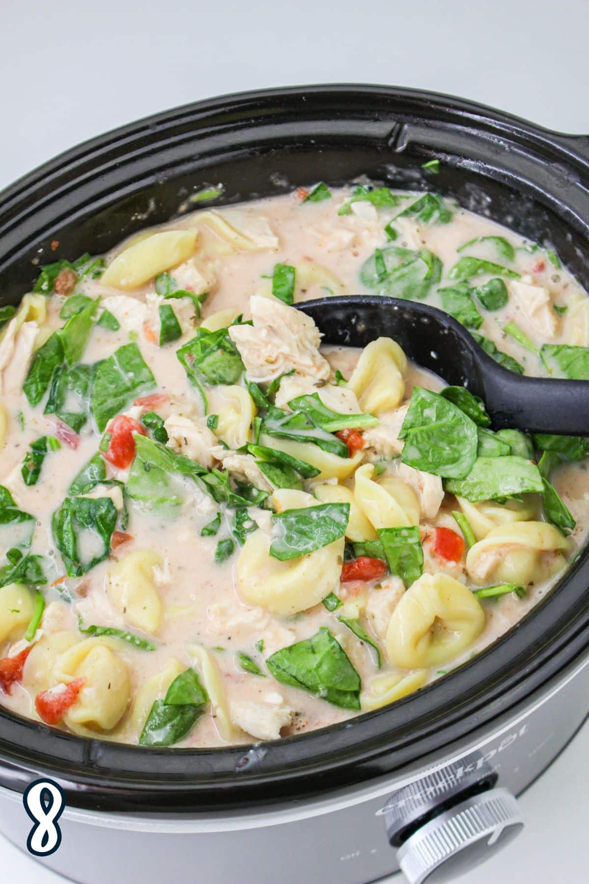 Creamy chicken tortellini soup with spinach and tomatoes in a slow cooker, with a black ladle for serving.