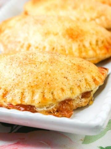 Close-up of golden-brown hand pies on a white rectangular plate, with flaky crusts and a sweet filling oozing out.