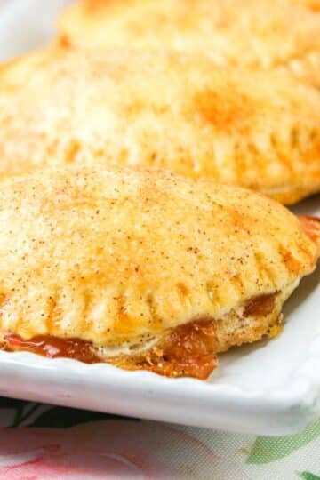 Close-up of golden-brown hand pies on a white rectangular plate, with flaky crusts and a sweet filling oozing out.